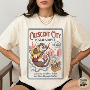 May include: Ivory-colored t-shirt featuring a vintage-style graphic. The graphic depicts an otter in a postal worker uniform with the text "CRESCENT CITY POSTAL SERVICE" and "Servicing the Istros River and Bone Quarter District". The shirt is made of Comfort Colors material.