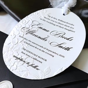 以下が含まれることがあります： エンボス加工された花の縁取りがある円形の白い結婚式の招待状。黒い文字で「Mr. & Mrs. William Brooks invite you to share their joy at the wedding of their daughter Emma Brooks to Alexander Smith, Saturday, the nineteenth of September, 2026 at half past five in the evening, The Edenvale Manor, San Jose, California, reception to follow」と書かれ、白いリボンで結ばれています。