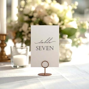 以下が含まれることがあります： テーブル番号「SEVEN」と筆記体の「table」が書かれた白いカード。カードは銅色の金属スタンドで支えられています。背景にはキャンドルとフラワーアレンジメントがあります。