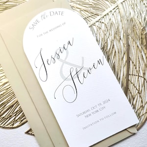 Pode incluir: Um convite de casamento branco com a parte superior arqueada, com os nomes "Jessica & Steven" em escrita elegante. O texto "Save the Date" e os detalhes do casamento são impressos em cinza claro. O convite é colocado em um envelope creme e uma folha de ouro decorativa.