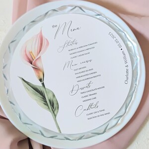 May include: A round white menu card featuring a pink calla lily illustration on the left side. The text reads The Menu, Starters, Main courses, Desserts, Cocktails, and Charlotte & William 05.05.2025. The card has a scalloped edge design.