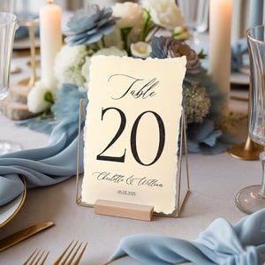 Peut inclure: Un carton de numéro de table 20 avec un cadre en métal doré et une base en bois. Le carton crème affiche le texte « Table 20 », les noms « Charlotte & William » et la date « 05.05.2025 ». La table est ornée de bougies et de compositions florales.