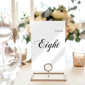 May include: A white table number card with the word "Eight" in elegant black script. The card is held in a gold-colored metal stand. The word "Table" is printed above the number. The setting includes a wine glass and candles.