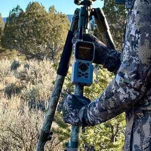 May include: A person wearing camouflage clothing uses a handheld electronic device mounted on a tripod in a wooded area.