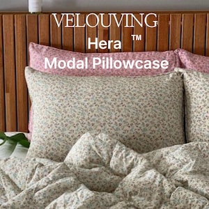 May include: Floral patterned pillowcase and bedding set. The pillowcase is cream-colored with a delicate floral print in shades of blue, purple, and brown. The text "VELOUVING Hera Modal Pillowcase" is visible. The bedding is on a wooden headboard.