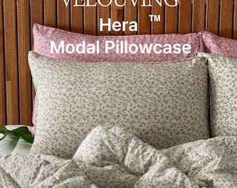May include: Floral patterned pillowcase and bedding set. The pillowcase is cream-colored with a delicate floral print in shades of blue, purple, and brown. The text "VELOUVING Hera Modal Pillowcase" is visible. The bedding is on a wooden headboard.