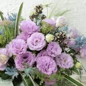May include: A floral arrangement featuring large, light purple flowers as the focal point. The bouquet includes blue, green, and white accents, with various types of foliage. The flowers are arranged in a white container against a textured white background.