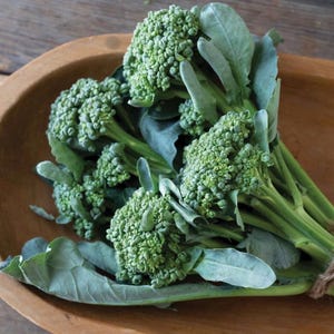 May include: Fresh green broccoli florets with large leaves, arranged in a wooden bowl. The broccoli has a vibrant green color, and the leaves are a muted green. The bowl is made of wood and has a natural finish.
