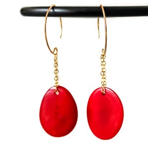 May include: A pair of gold-toned earrings with a chain and a large, oval, red stone. The earrings are hanging from a black bar against a white background. The earrings are a simple, elegant design.
