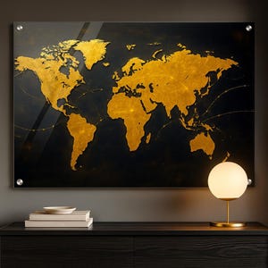 Könnte beinhalten: Ein rechteckiges Wandbild mit einer Weltkarte, auf der goldene Kontinente vor einem dunklen Hintergrund dargestellt sind. Die Karte ist auf einer transparenten Oberfläche mit silbernen Beschlägen montiert. Eine goldfarbene Lampe mit kugelförmigem Schirm steht auf einer dunklen Holzoberfläche.