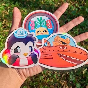 Peut inclure: Trois autocollants colorés représentant des personnages de dessins animés. L'un montre un pingouin avec un chapeau de marin et une bouée de sauvetage. Un autre représente un chat dans un sous-marin orange. Le troisième a un visage souriant avec des plantes aquatiques.