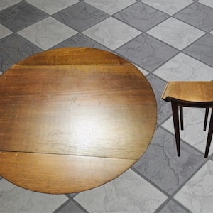 May include: A round wooden table with a smooth, medium-brown finish and a visible seam across the surface, accompanied by a small, matching wooden stool featuring four tapered legs and a curved seat top, both displaying a consistent wood grain.