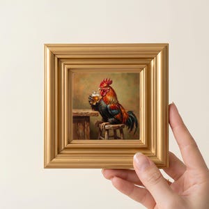 May include: A square oil painting in a thick gold frame depicting a rooster with red, black, and orange feathers sitting on a wooden stool. The rooster is holding a small glass of amber liquid in its wing, positioned next to a wooden table.