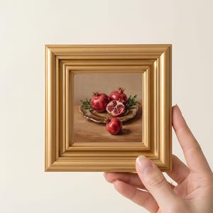 May include: A small square oil painting in a thick gold frame depicting a still life of pomegranates. The artwork shows three whole red pomegranates and one halved pomegranate with visible seeds, arranged on a shallow plate against a neutral background.
