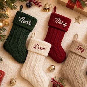 Könnte beinhalten: Vier Weihnachtsstrümpfe aus Zopfstrick in Dunkelgrün, Tiefrot, Creme und Beige. Jeder Strumpf hat einen umgeschlagenen Bund, der mit den Namen Noah, Merry, Liam und Joy in Schreibschrift bestickt ist, sowie eine Schlaufe zum Aufhängen.