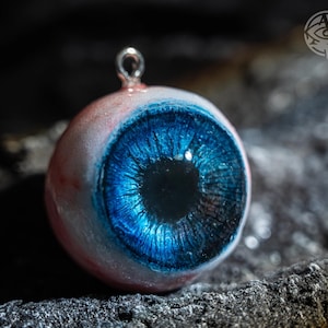 Puede incluir: Un colgante detallado con forma de ojo humano. El ojo presenta un iris azul vibrante con detalles intrincados, rodeado por una esclerótica blanca con venas rojas. Un lazo plateado está unido en la parte superior para colgar.