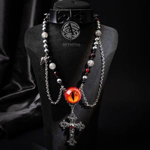 May include: A black choker necklace with silver studs and a pendant necklace. The pendant features a red eye design, a silver cross, and a chain with beads. The necklace is displayed on a black mannequin. The brand name "AETHERIA" is visible.