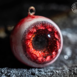 Puede incluir: Primer plano de un colgante con forma de ojo humano. El ojo tiene un iris rojo y una pupila negra. El ojo está sobre un fondo oscuro y tiene un lazo dorado en la parte superior.
