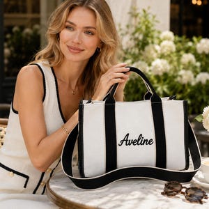 May include: A white canvas tote bag with black trim and a black shoulder strap. The bag features the name Aveline embroidered in black script on the front center. It has two black top handles and a rectangular shape with a structured design.