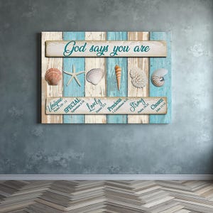 Peut inclure: Une œuvre d'art murale en bois sur le thème de la plage. L'art présente un fond en bois bleu clair et blanc avec des coquillages et une étoile de mer. Le texte "Dieu dit que tu es" est en haut, et d'autres mots sont en dessous.