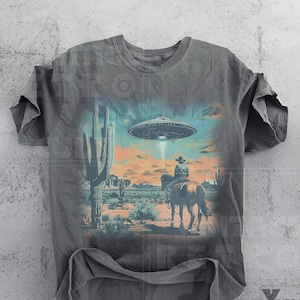 Pu&ograve; includere: Una maglietta grigia con una stampa grafica di un cowboy a cavallo in un paesaggio desertico con un UFO nel cielo. La grafica &egrave; in stile vintage.