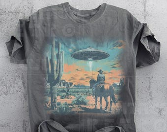 May include: A gray t-shirt with a graphic print of a cowboy riding a horse in a desert landscape with a UFO in the sky. The graphic is in a vintage style.
