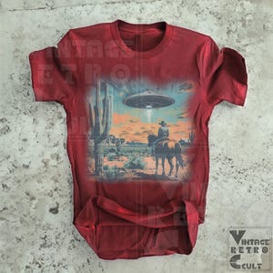 Pu&ograve; includere: Una maglietta rossa con una stampa grafica di un cowboy a cavallo in un paesaggio desertico con un UFO nel cielo. La grafica &egrave; in tonalit&agrave; di blu, verde e marrone.
