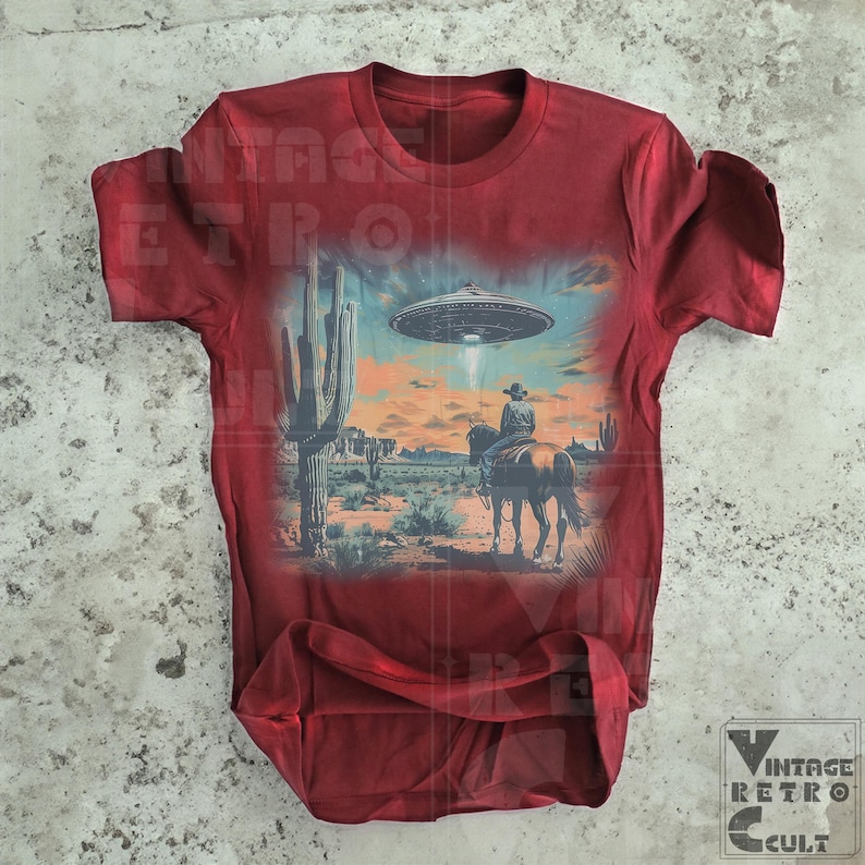 Pu&ograve; includere: Una maglietta rossa con una stampa grafica di un cowboy a cavallo in un paesaggio desertico con un UFO nel cielo. La grafica &egrave; in tonalit&agrave; di blu, verde e marrone.