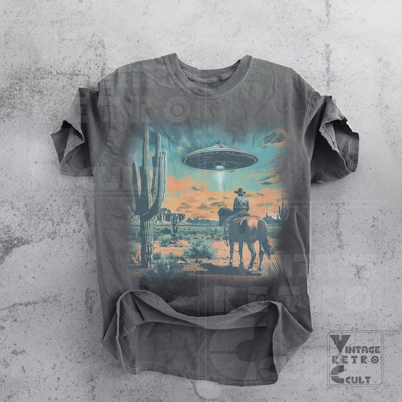 Pu&ograve; includere: Una maglietta grigia con una stampa grafica di un cowboy a cavallo in un paesaggio desertico con un UFO nel cielo. La grafica &egrave; in stile vintage.
