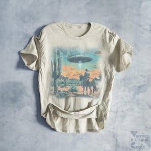 Pu&ograve; includere: Una t-shirt beige con una stampa grafica di un cowboy a cavallo in un paesaggio desertico con un UFO nel cielo. La grafica &egrave; in tonalit&agrave; di blu, verde e arancione.