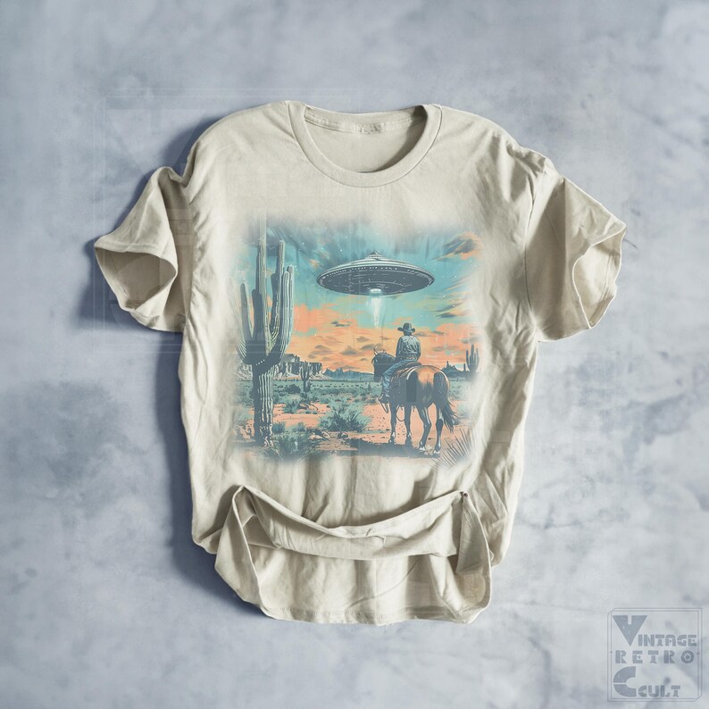 Pu&ograve; includere: Una t-shirt beige con una stampa grafica di un cowboy a cavallo in un paesaggio desertico con un UFO nel cielo. La grafica &egrave; in tonalit&agrave; di blu, verde e arancione.