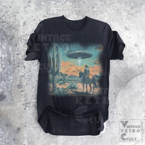 Pu&ograve; includere: T-shirt nero con una grafica vintage di un cowboy a cavallo davanti a un paesaggio desertico con un UFO nel cielo. Il testo "Vintage Retro Cult" &egrave; stampato sulla maglietta.