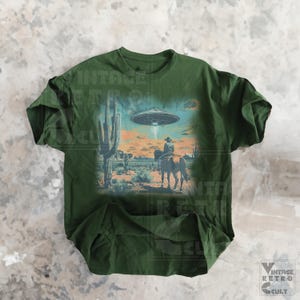 Pu&ograve; includere: Una maglietta verde con una stampa grafica di un cowboy a cavallo in un paesaggio desertico con un UFO nel cielo. La grafica &egrave; in stile vintage.