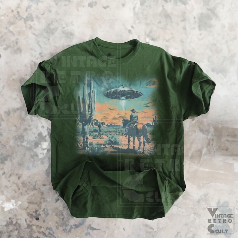 Pu&ograve; includere: Una maglietta verde con una stampa grafica di un cowboy a cavallo in un paesaggio desertico con un UFO nel cielo. La grafica &egrave; in stile vintage.