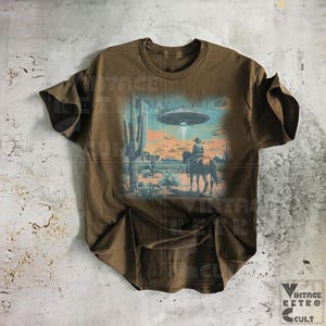 Pu&ograve; includere: Una maglietta marrone con una stampa grafica vintage di un cowboy a cavallo in un paesaggio desertico con un UFO nel cielo. La grafica &egrave; in tonalit&agrave; di blu, verde e arancione.