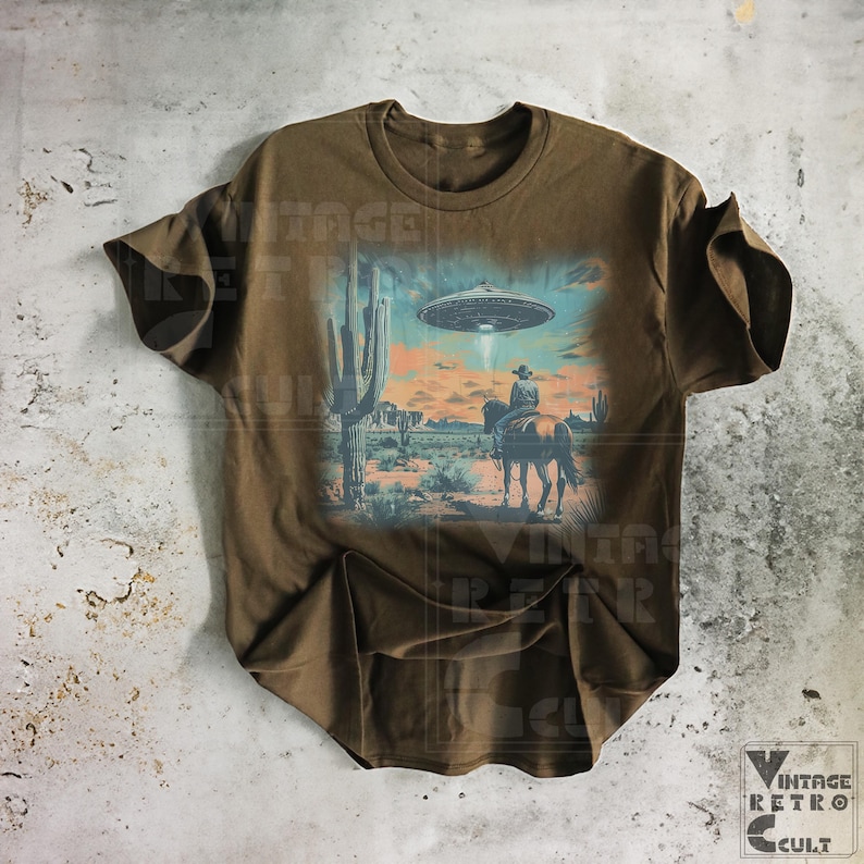 Pu&ograve; includere: Una maglietta marrone con una stampa grafica vintage di un cowboy a cavallo in un paesaggio desertico con un UFO nel cielo. La grafica &egrave; in tonalit&agrave; di blu, verde e arancione.