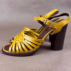 May include: Mustard yellow sandals with a woven strap design and a buckle closure. The sandals have a dark brown sole and a wooden heel that is approximately 3 inches (7.6 cm) high.