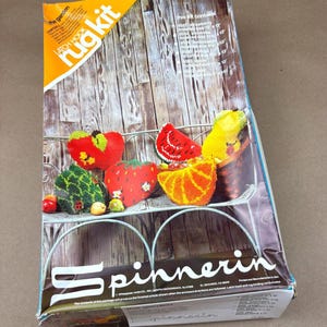 May include: A vintage "Spinnerin" latch hook rug kit featuring a wooden background and a metal shelf with colorful fruit designs. The kit includes a strawberry, watermelon, orange, and other fruit patterns. The box reads "The Garden".