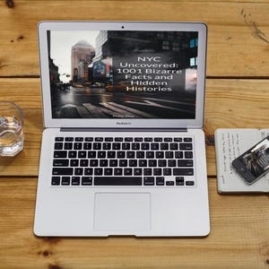 May include: A silver MacBook Air laptop displaying the text "NYC Uncovered: 1001 Bizarre Facts and Hidden Histories" on the screen. A glass of water, a smartphone, and a notebook are on a wooden surface.