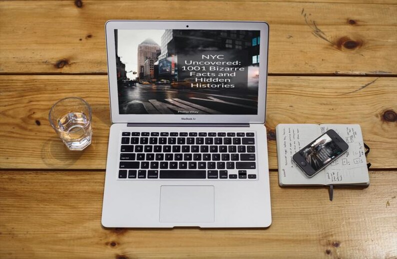 May include: A silver MacBook Air laptop displaying the text "NYC Uncovered: 1001 Bizarre Facts and Hidden Histories" on the screen. A glass of water, a smartphone, and a notebook are on a wooden surface.