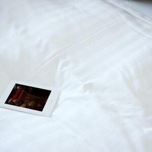 May include: A white tablet displaying a dark image with red text, resting on a white bedsheet. The bedsheet has subtle vertical lines. A patterned pillow is visible in the background.