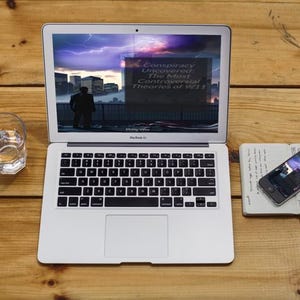 Peut inclure: Un ordinateur portable MacBook Air argent&eacute; affichant du texte sur les th&eacute;ories du complot du 11/09, un verre d'eau, un smartphone et un carnet sur une surface en bois. L'&eacute;cran de l'ordinateur portable montre une silhouette.