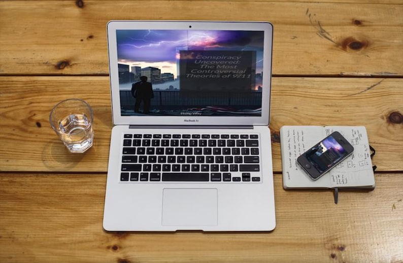 Peut inclure: Un ordinateur portable MacBook Air argent&eacute; affichant du texte sur les th&eacute;ories du complot du 11/09, un verre d'eau, un smartphone et un carnet sur une surface en bois. L'&eacute;cran de l'ordinateur portable montre une silhouette.