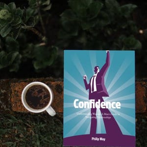 以下が含まれることがあります： 「Confidence」というタイトルの本。ターコイズ色の表紙にスーツ姿の男性のグラフィック。本の隣には、濃いコーヒーが入った白いコーヒーカップがあります。背景には緑とレンガの壁があります。