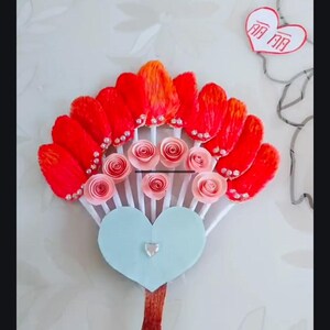 May include: A red and orange paper fan with pink paper roses and white sticks. The fan is attached to a blue heart-shaped base with a silver rhinestone. The base is attached to a red stick.