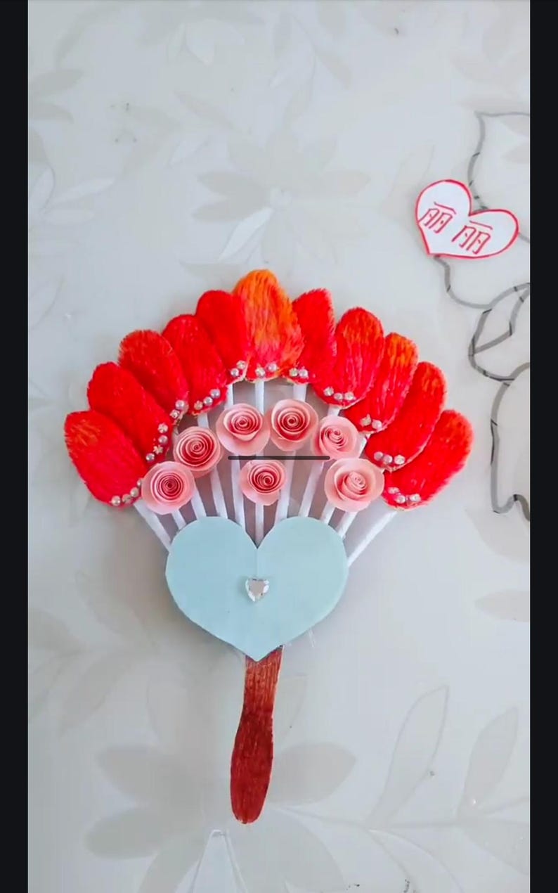 May include: A red and orange paper fan with pink paper roses and white sticks. The fan is attached to a blue heart-shaped base with a silver rhinestone. The base is attached to a red stick.
