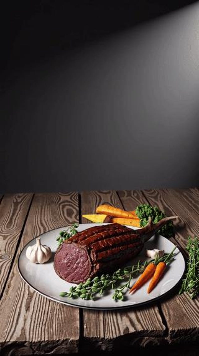 May include: A cooked ribeye steak with the bone in, presented on a white plate with fresh vegetables. The plate includes carrots, garlic, and herbs. The steak is a deep brown colour with a pink interior, set on a wooden table.