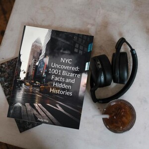 May include: A book titled "NYC Uncovered: 1001 Bizarre Facts and Hidden Histories" rests on a table next to a pair of black headphones and a glass of iced coffee. The book cover features a city street scene. A small, patterned card is also visible.