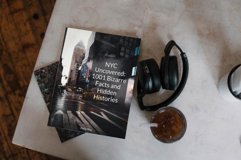 May include: A book titled "NYC Uncovered: 1001 Bizarre Facts and Hidden Histories" rests on a table next to a pair of black headphones and a glass of iced coffee. The book cover features a city street scene. A small, patterned card is also visible.