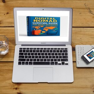 Op de afbeelding: Een laptop toont de boekomslag "DIGITAL NOMAD SURVIVAL GUIDE FOR BROKE BEGINNERS". Een smartphone en notitieboek liggen ernaast. Een glas water staat links op een houten oppervlak. De sc&egrave;ne suggereert een werkplek.